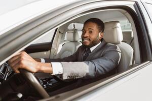 A black man wearing a suit is driving a car on a sunny day. He is smiling and appears to be enjoying the drive. The car is modern and sleek, with a white exterior and a luxurious interior. photo