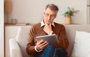 Serious Elderly Gentleman Using Tablet Computer Browsing Internet Sitting On Couch At Home. Retired Life. Selective Focus photo