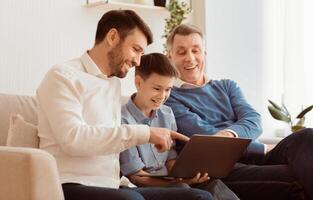 Boy Using Laptop Sitting Between Father And Grandpa Doing Homework Online On Sofa At Home. Selective Focus photo