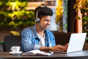 A young black guy is seated in a coffee shop, focused on using his laptop. He is typing on the keyboard, surrounded by tables and chairs. The background shows other customers and baristas. photo