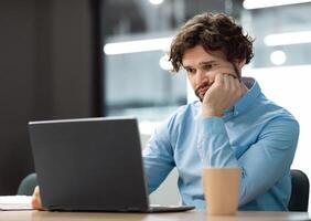 Portrait of sad bored business man sitting at desk using pc, leaning head on hand looking at screen. Upset stressed guy suffering job problems, reading bad negative news at office, free copy space photo