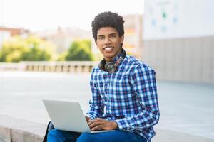 A teen black guy sits outside with headphones around his neck, using a laptop computer. He is smiling and appears to be focused on his work. The image is taken on a sunny day photo