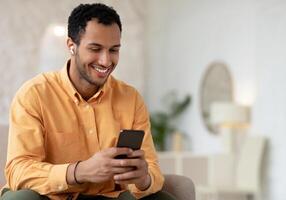 Smiling Arabic Male Listening To Music On Mobile Phone Wearing Wireless Headphones Sitting On Couch Relaxing At Home. Male Enjoying Favorite Playlist Using Musical Application, Free Copy Space photo