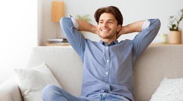 A man is sitting on a white couch, relaxed and smiling, with his arms behind his head. photo