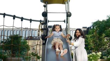 Young girl enjoys sliding down playground slide while mother supervises in city park video