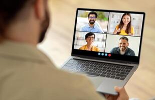 A man sitting at a table is using a laptop, which displays a group of people on the screen. He appears engaged in a virtual meeting or conference with his colleagues. photo