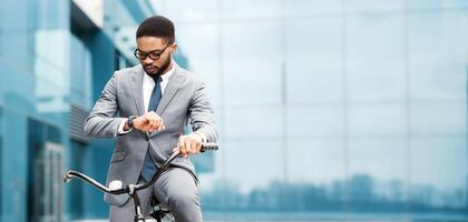 African American businessman in a suit is on a bicycle, looking at his watch in a city setting. The scene is busy and modern, emphasizing efficiency and time management in a professional context. photo