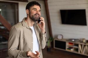 A man is engaged in a phone call while standing in a modern living room, holding a cell phone to his ear. The room is tastefully decorated with a sofa, coffee table, and plants, copy space photo