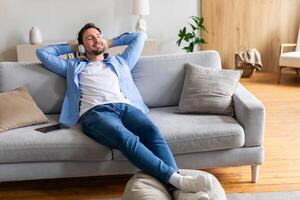 A man is comfortably seated on a couch in his living room, appearing relaxed and at ease. The room is well-lit with natural light, and the man seems to be enjoying a moment of rest and relaxation. photo