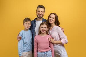 A family of four, consisting of two adults and two children, stands together in front of a vibrant yellow background, smiling and posing for a photograph, with the parents holding the children hands photo