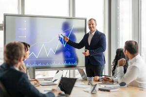 A businessman in a suit stands in front of a large screen, pointing to an upward trending graph. He is presenting to a group of colleagues who are seated at a conference table, listening attentively photo