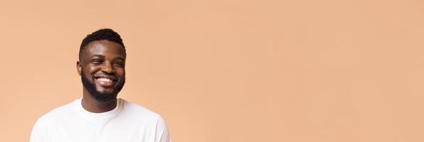 A young black man with a beard, wearing a plain white t-shirt, smiles broadly against a peach-colored background. He is looking directly at the camera, with a natural and warm expression, copy space photo