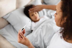 A caring African American mother is gently placing her hand on her child forehead to feel for a fever while holding a digital thermometer. The child appears to be resting in bed photo