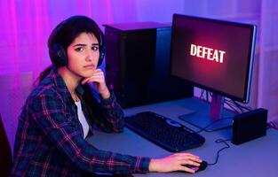 A young woman, wearing headphones, sits at a computer desk with a serious expression on her face. The word DEFEAT is displayed on the computer screen, indicating that she has lost a game photo
