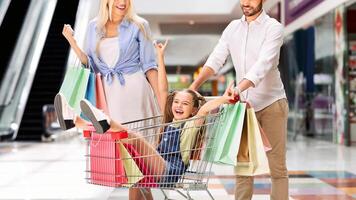 A young girl sits in a shopping cart, being pushed by her parents. The girl is smiling and laughing as she holds onto the handle of the cart. Her parents are also smiling and look happy photo