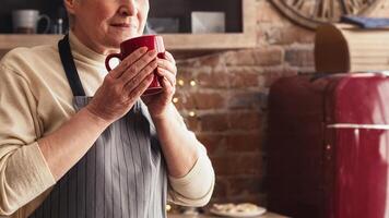 A woman wearing an apron holds a red mug in her hands, smiling and taking a moment to enjoy a warm drink. She is in a cozy kitchen, with brick walls and a red refrigerator in the background photo