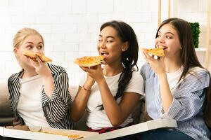 Three young girls are seated on a couch, enjoying slices of pizza. The girls are casually dressed and appear to be having a relaxed and enjoyable time together. photo
