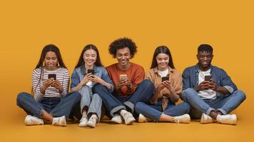 This image features a diverse group of five young adults, all smiling and looking down at their smartphones. They are sitting cross-legged on a yellow background photo