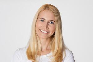 The image is a portrait of a middle aged woman with long blonde hair. She is smiling and looking directly at the camera. She is wearing a white shirt and standing in front of a white background photo
