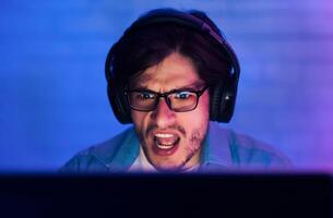A young man is shown intensely focused on a computer screen, wearing headphones and glasses. His mouth is open in a shout, likely reacting to an exciting moment in a game. photo