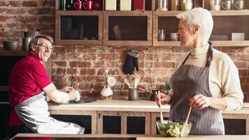 A senior couple is cooking together in a modern kitchen. The man is chopping vegetables, while the woman is standing by the stove. They are both wearing aprons and smiling. photo