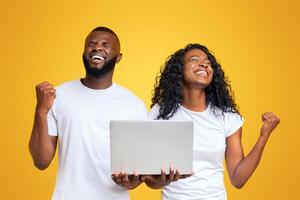 Extremely happy black couple with closed eyes celebrating victory, clenching fists, holding laptop photo