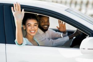 African American young woman and man are smiling and waving from the window of a white car. They appear to be happy and enjoying their time together. photo