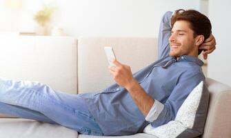 A man is relaxing on a couch, using his smartphone. He is smiling and appears to be enjoying himself. photo