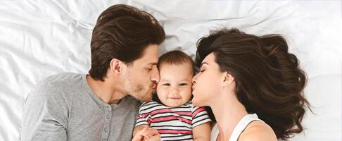 A father and mother lay on a white bed and kiss their baby, who is lying between them. The father is on the left side of the image, and the mother is on the right, top view, panorama photo