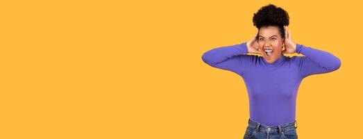 A young woman with curly dark hair is standing in front of a bright yellow background. She has her hands over her ears and is screaming with her mouth open, panorama with copy space photo
