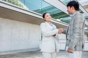 Two professionals man and woman engaging in a friendly handshake outside a contemporary building. They appear to be discussing business matters amidst a clear sky and a sleek architectural backdrop. photo