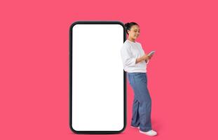 Asian woman wearing a white sweater and blue jeans stands beside a large smartphone with a blank white screen. The background is a vibrant pink color. She holds another smartphone photo