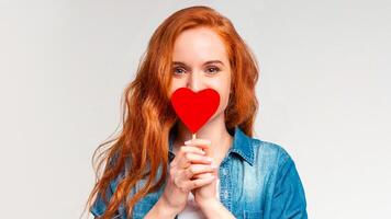 A woman is holding a red heart in front of her face, covering most of her features. She appears to be expressing love or affection towards the viewer, with a soft smile on her lips. photo