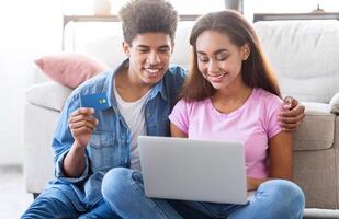 Black teen couple sits on the floor in front of a couch, using a laptop to shop online. The girl is typing on the laptop, while the guy holds a credit card and looks at the screen. photo