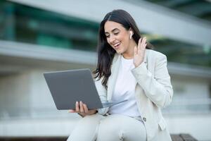 A woman in a white blazer and pants sits outside in front of a building, using her laptop for a call. She is smiling and waving at the screen, likely participating in a business meeting photo