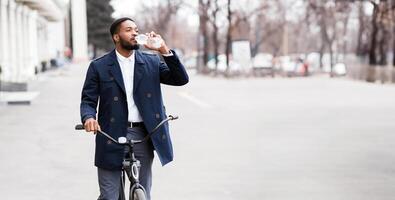 An executive black guy is cycling to the office through downtown, drinking water. The mood is lively and productive, with an urban backdrop highlighting a modern and active commute. photo