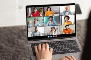 A woman is typing on a laptop computer, participating in a conference with eight other people. The screen is displaying nine different feeds of the participants photo