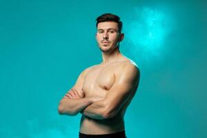 A confident young man stands shirtless with arms crossed, looking directly at the camera. The background is a vibrant blue, accentuated by fog effects, creating a dramatic and striking atmosphere. photo