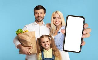 A happy family of three stands in front of a blue background. The mother holds a smartphone with a blank screen towards the camera. The father holds a paper bag full of groceries. photo