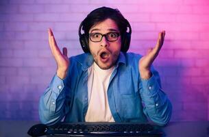 This image features a young man, wearing headphones, sitting in front of a computer and looking shocked and surprised. He is wearing a blue shirt and appears to be playing a game. photo