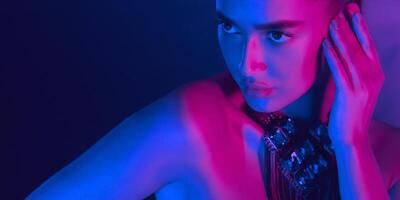 A woman is bathed in blue and pink neon lighting at night. She is touching her face with one hand while gazing intently into the distance, creating a dramatic and captivating visual effect. photo