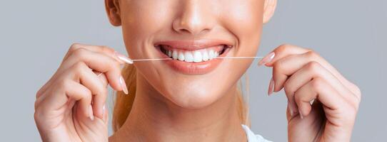 A close-up image of a woman smiling as she uses white floss to clean her teeth. She is holding the floss with both hands, and her teeth are visible in the center of the image. photo