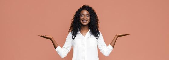 Smiling African American woman in white shirt extending her hands with palms up, making choice isolated on peach studio background photo
