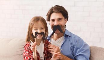 Beautiful funny little girl and daddy have mustaches on sticks, posing at home. Family holidays and togetherness concept, copy space photo