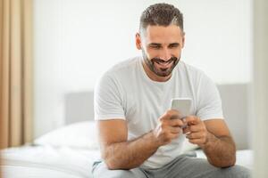 A man in a white shirt sits on a white bed, smiling as he looks at his smartphone. He is sitting with his legs crossed and his arms are resting on his knees. The bed is made with a white sheet. photo