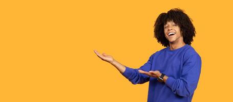 A cheerful young man with curly hair is enthusiastically gesturing with both hands against a solid yellow background, panorama with copy space photo