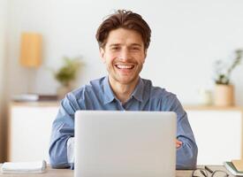 A smiling man with crossed arms sits at a desk in an office, working on a laptop. photo