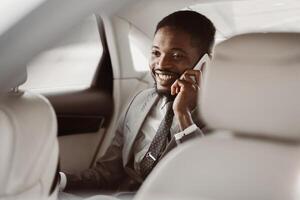 Successful African American businessman, wearing a suit and tie, sits in the backseat of a car and talks on a phone while smiling. photo