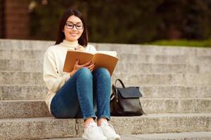 Asian woman student is seated on a set of steps, engrossed in reading a book. She is holding the book in her hands and appears focused on the pages. photo