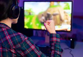 A young gamer, wearing headphones and a plaid shirt, raises their fist in celebration, seemingly victorious in a game displayed on the computer monitor. photo